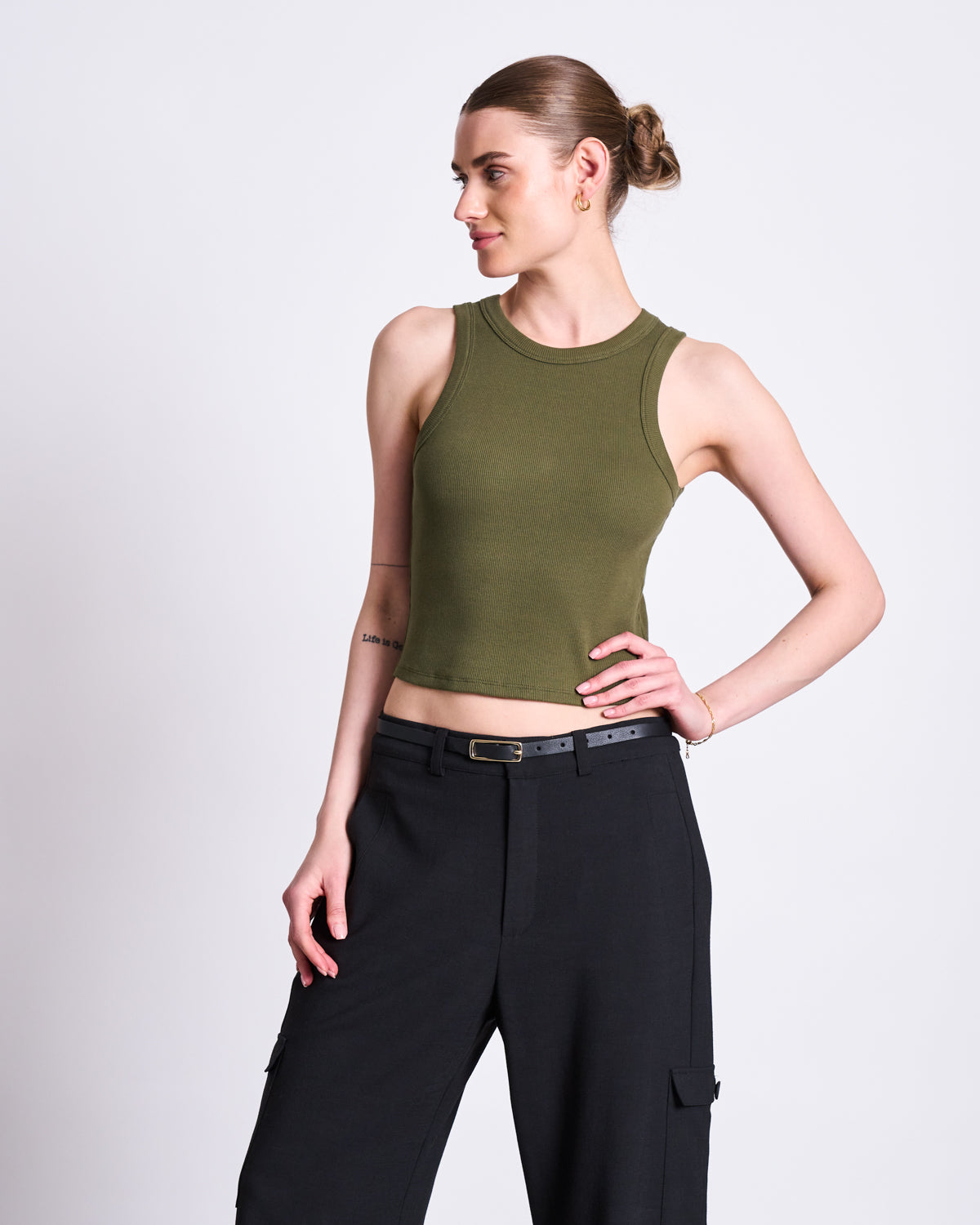 A woman with her hair in a bun wears the jan-n-june ORGANIC COTTON TANK TOP PORTO DARK OLIVE GOTS and black belted cargo pants, standing against a plain white background and looking to the side with a relaxed pose.