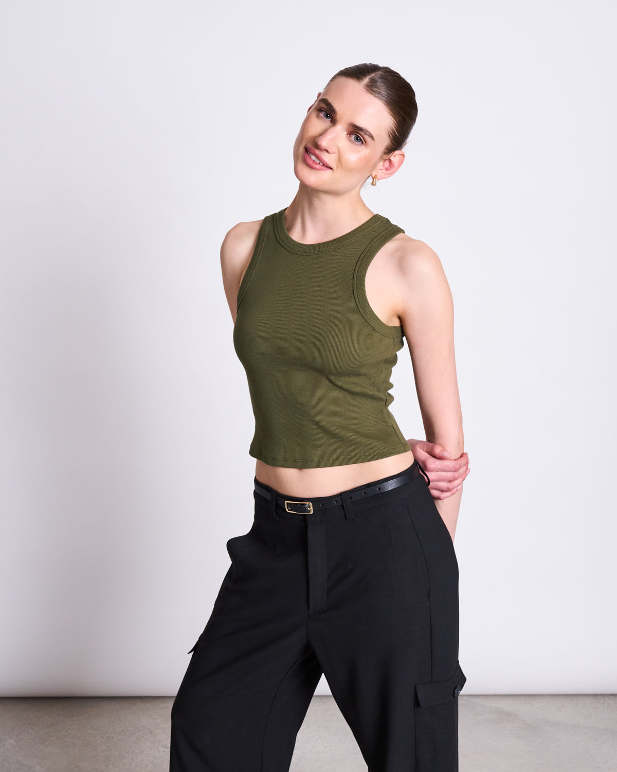 A woman with light skin and brown hair in a low bun wears the jan-n-june ORGANIC COTTON TANK TOP PORTO DARK OLIVE GOTS and black pants, standing against a plain light background, tilting her head and smiling softly with arms behind her back.