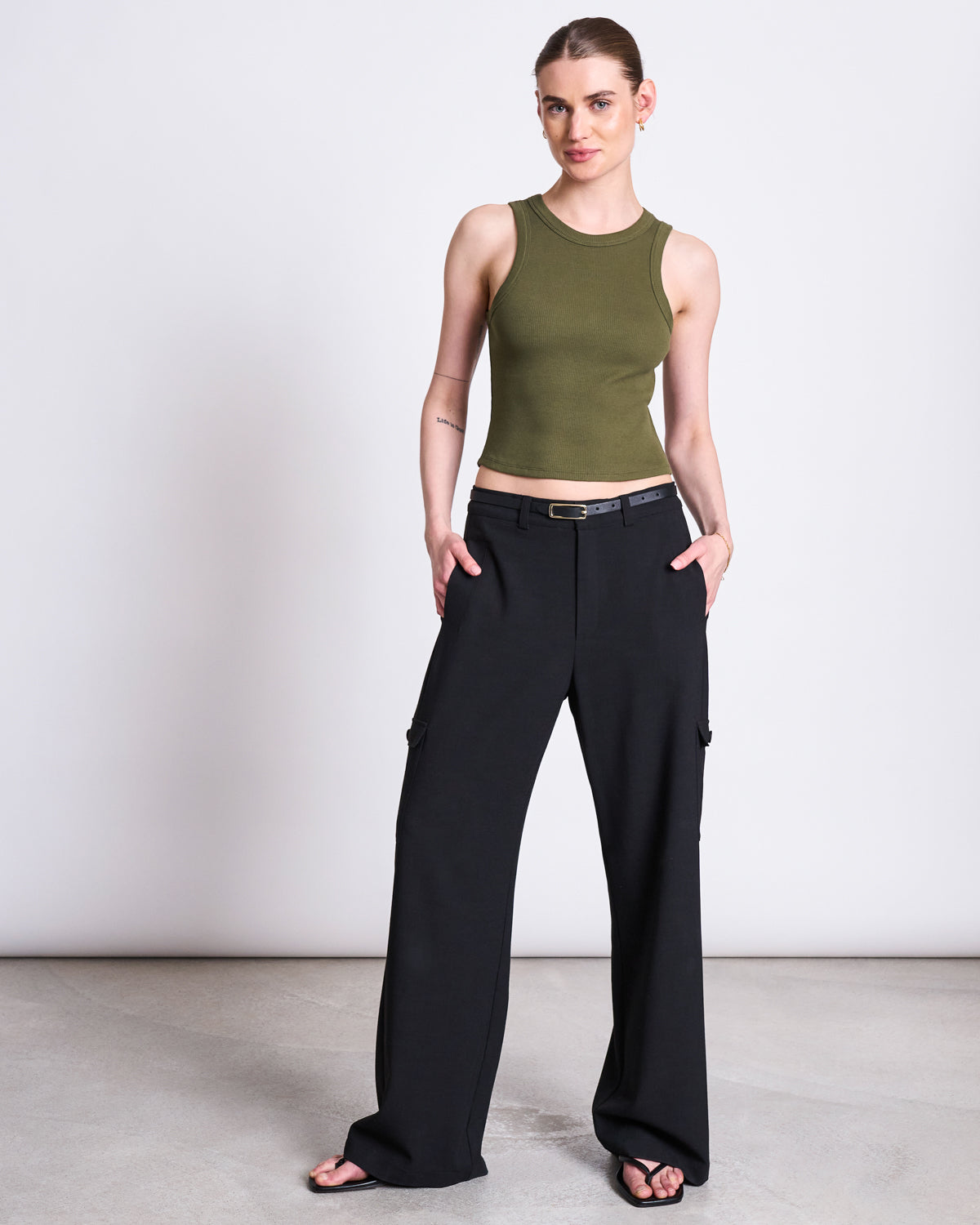 A woman stands against a plain background wearing a jan-n-june ORGANIC COTTON TANK TOP PORTO DARK OLIVE GOTS and black wide-leg cargo pants with a belt, her hair pulled back and hands in pockets.