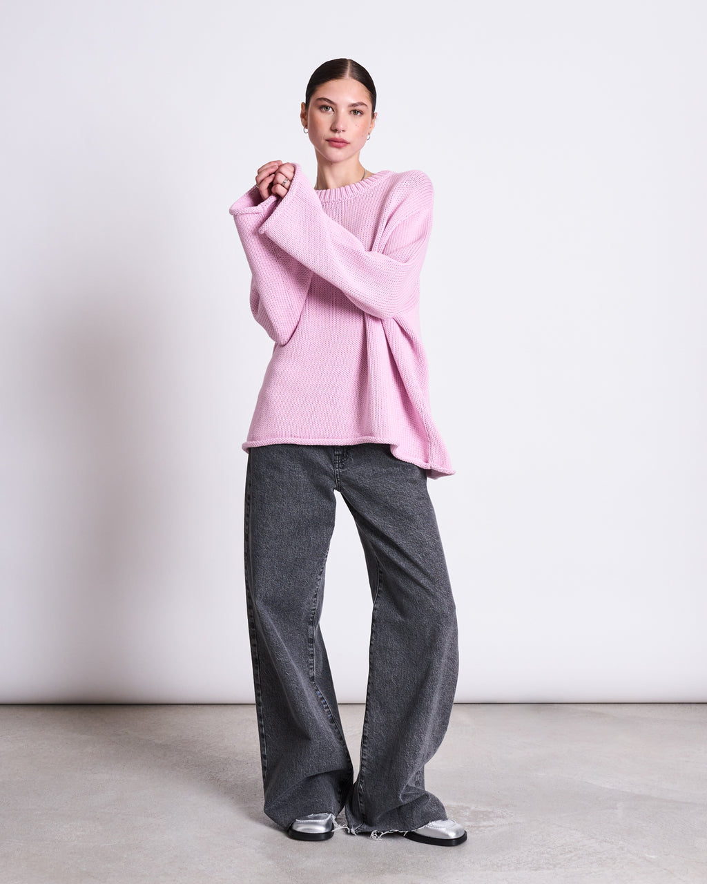 A woman stands against a plain backdrop wearing the jan-n-june JUMPER SONA LIGHT ORCHID GOTS, an oversized light pink knit sweater with extra-long sleeves, paired with wide-leg gray jeans and silver sneakers. Her hair is tied back.