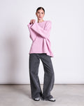 A woman stands against a plain backdrop wearing the jan-n-june JUMPER SONA LIGHT ORCHID GOTS, an oversized light pink knit sweater with extra-long sleeves, paired with wide-leg gray jeans and silver sneakers. Her hair is tied back.