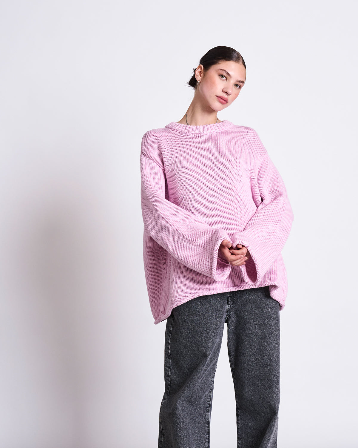 A person with dark hair pulled back stands against a plain light background, wearing jan-n-june’s oversized JUMPER SONA LIGHT ORCHID GOTS sweater in light pink and dark gray jeans, looking slightly to the side with hands clasped.