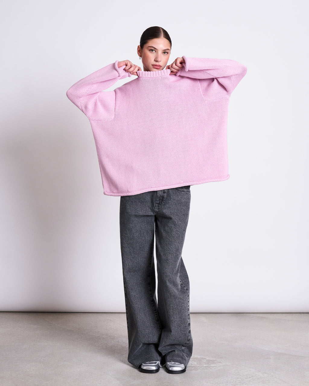 A woman poses against a plain background wearing the jan-n-june JUMPER SONA LIGHT ORCHID GOTS, an oversized light pink organic cotton sweater with wide sleeves, styled with dark gray wide-leg jeans and sneakers, holding the collar over her face.