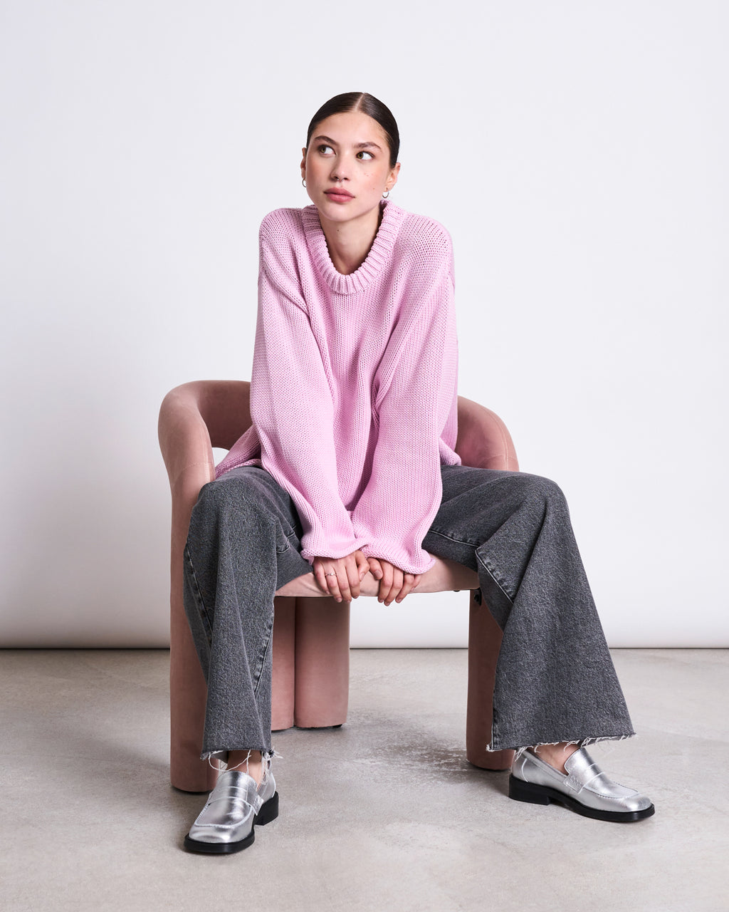 A woman sits on a pink chair, wearing the jan-n-june JUMPER SONA LIGHT ORCHID GOTS, paired with wide-leg gray jeans and silver loafers. She looks to her right with hands in front of her against a minimal background.