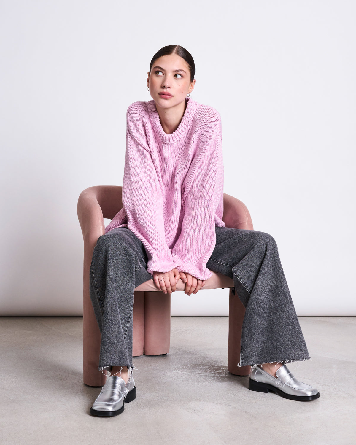 A woman sits on a pink chair, wearing the jan-n-june JUMPER SONA LIGHT ORCHID GOTS, paired with wide-leg gray jeans and silver loafers. She looks to her right with hands in front of her against a minimal background.
