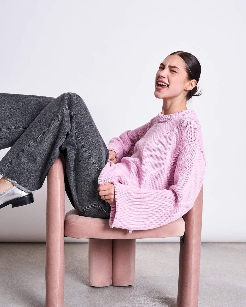 A woman wearing the jan-n-june JUMPER SONA LIGHT ORCHID GOTS sits sideways on a pink chair in gray jeans, laughing with her head tilted back and eyes closed.