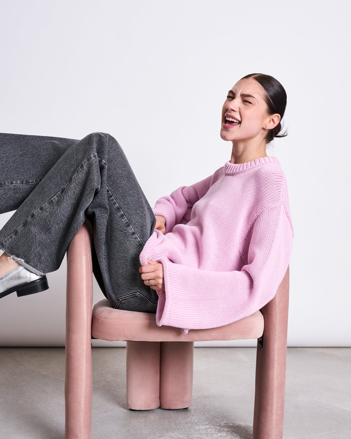 A woman wearing the jan-n-june JUMPER SONA LIGHT ORCHID GOTS sits sideways on a pink chair in gray jeans, laughing with her head tilted back and eyes closed.
