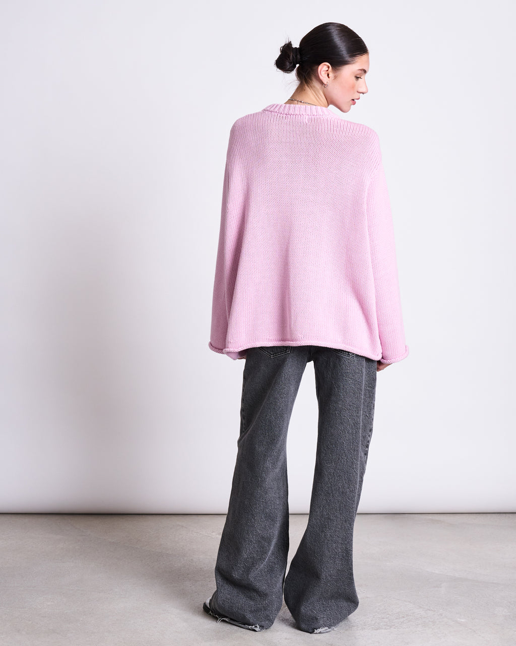 A person with dark hair in a bun wears the jan-n-june JUMPER SONA LIGHT ORCHID GOTS, an oversized organic cotton knit sweater in pink, styled with wide-leg gray jeans, standing on a concrete floor against a plain light background.