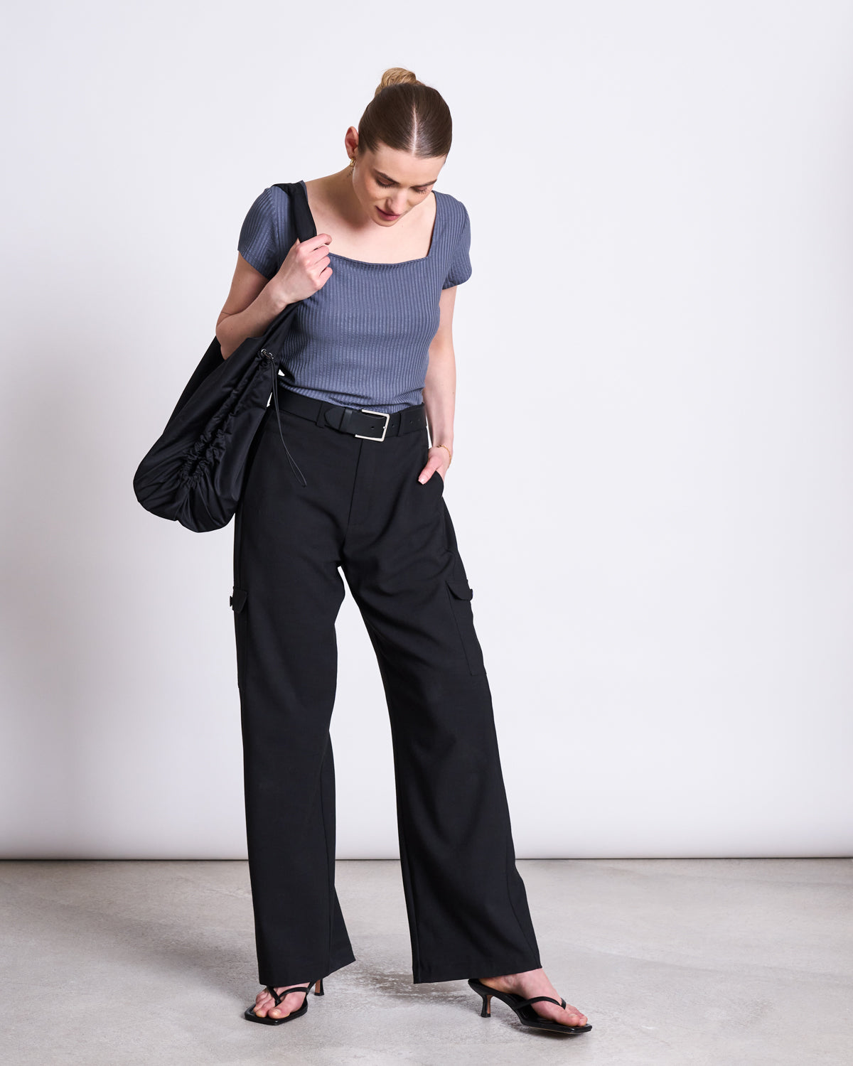 A woman with her hair tied back stands against a plain background, looking down. She wears the jan-n-june T-SHIRT ARIMA TURBULENCE, black wide-leg trousers, heeled sandals, and carries a large black shoulder bag.