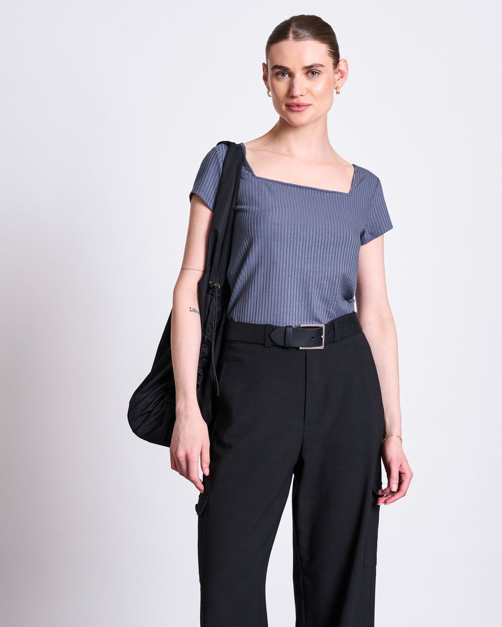 A woman with her hair pulled back wears the jan-n-june T-SHIRT ARIMA TURBULENCE, a gray square neck top in soft ribbed fabric, tucked into belted high-waisted black pants. She carries a black shoulder bag against a plain light background.