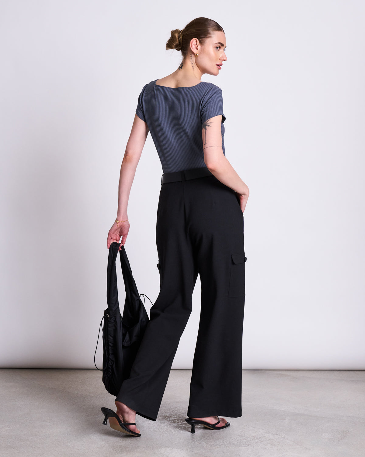 A woman with her hair in a bun, facing away, wears the gray jan-n-june T-SHIRT ARIMA TURBULENCE with black wide-leg pants and heeled sandals, holding a black bag against a white background.