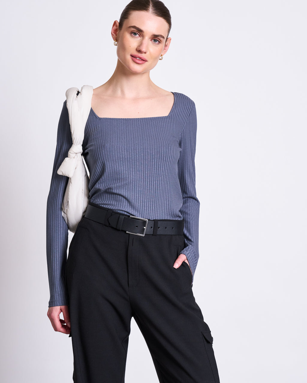 A woman with her hair tied back stands against a plain background wearing the jan-n-june SQUARENECK LONGSLEEVE FRIA TURBULENCE, black belted pants, and a light sweater over her shoulder, with one hand in her pocket.