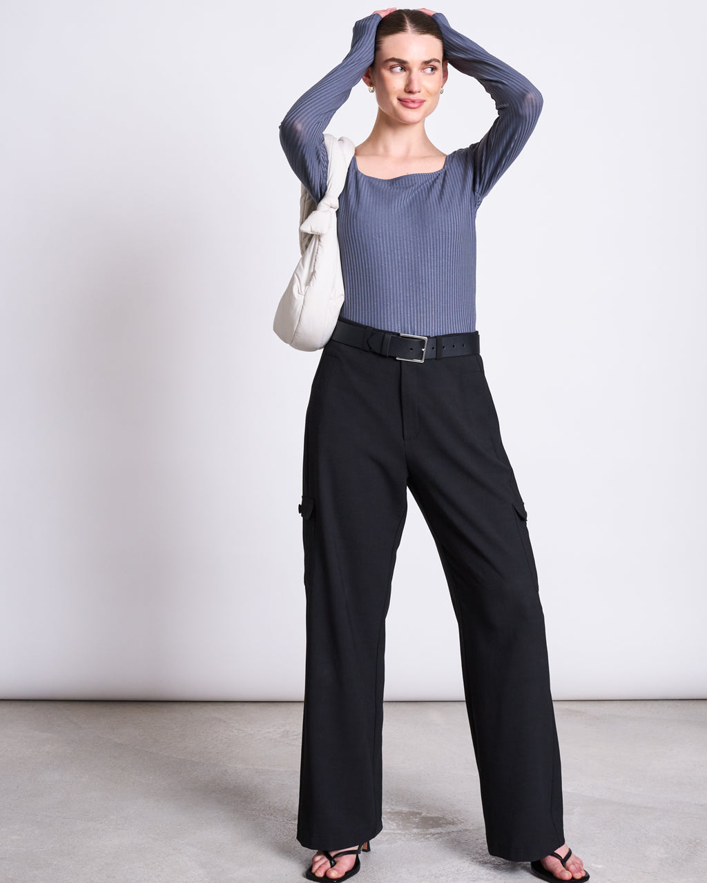 A woman stands against a plain background, wearing jan-n-june’s CARGO PANTS FANO BLACK with a belt, paired with a ribbed blue long-sleeve top, black heeled sandals, and a white shoulder bag. She rests her hands on her head and smiles softly.