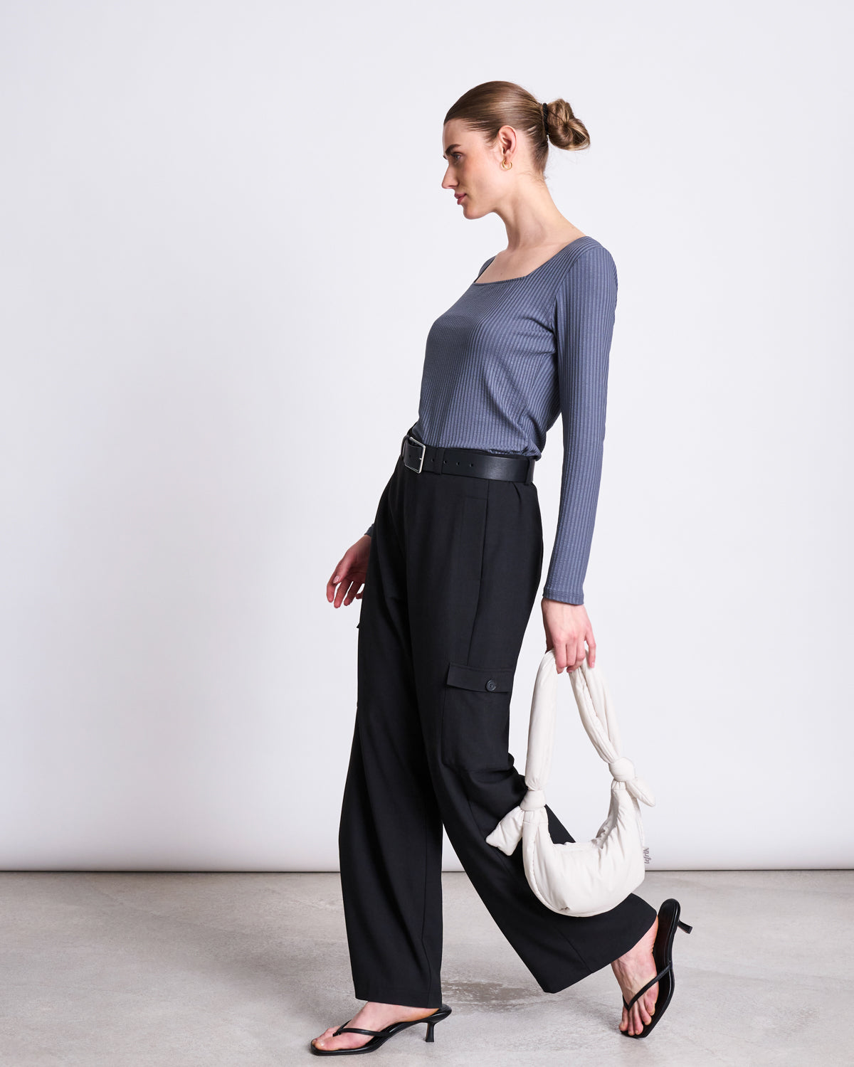 A woman with her hair in a bun wears the jan-n-june SQUARENECK LONGSLEEVE FRIA TURBULENCE ribbed blue top, black wide-leg trousers, and black sandals, carrying a white knotted handbag against a plain light background.
