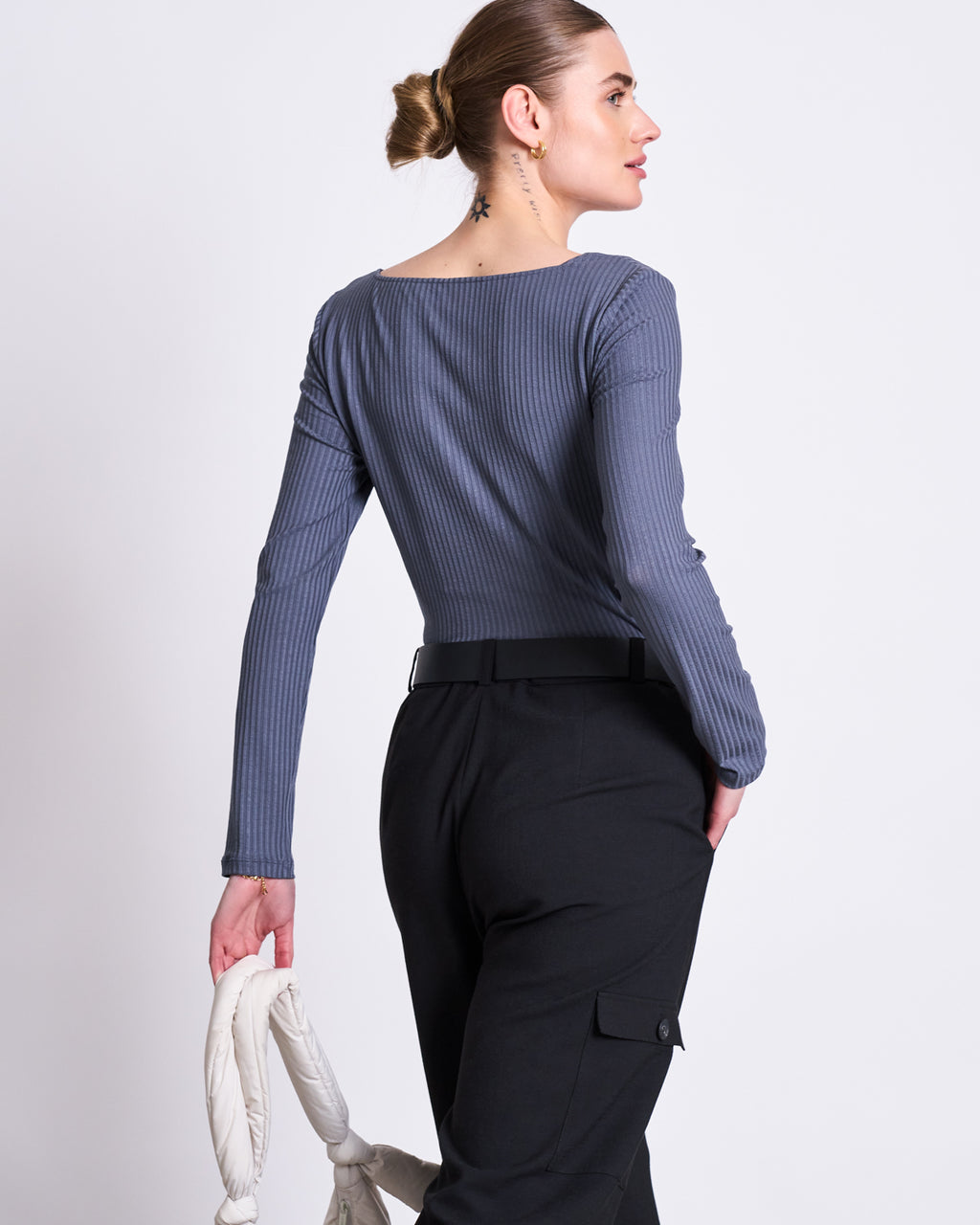 A woman with her hair in a bun wears jan-n-junes CARGO PANTS FANO BLACK, paired with a long-sleeve, ribbed grey top made from recycled polyester. She holds a white rope bag and faces slightly away from the camera against a plain background.