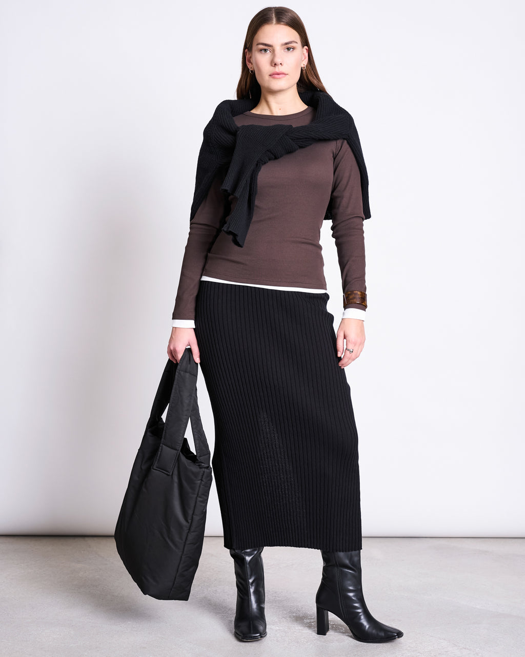 A woman stands against a plain background wearing the jan-n-june MIDI KNIT SKIRT EBBA BLACK GOTS, paired with a brown long-sleeve top, black heeled boots, a black sweater over her shoulders, and holding a large black tote bag.
