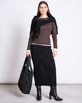 A woman stands against a plain background wearing the jan-n-june MIDI KNIT SKIRT EBBA BLACK GOTS, paired with a brown long-sleeve top, black heeled boots, a black sweater over her shoulders, and holding a large black tote bag.