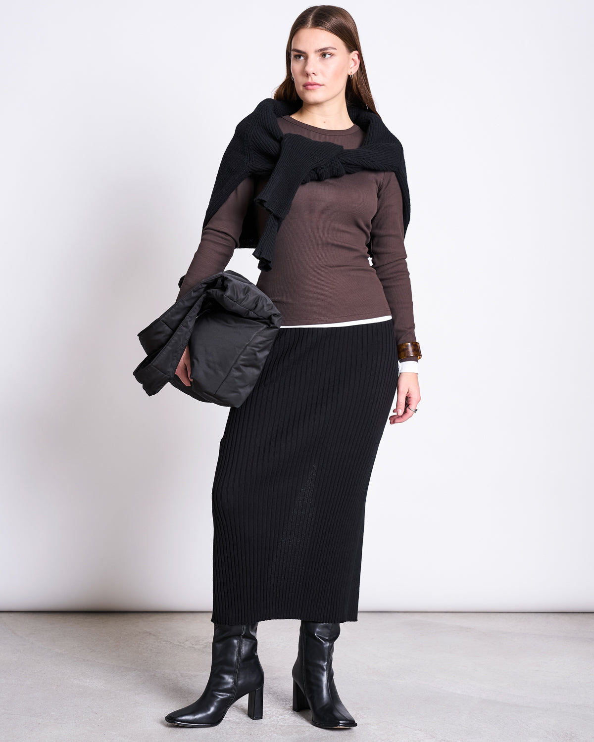 A woman stands against a plain background wearing the jan-n-june MIDI KNIT SKIRT EBBA BLACK GOTS with a brown long-sleeve top, black heeled boots, a black sweater over her shoulders, and holding a puffy black jacket.