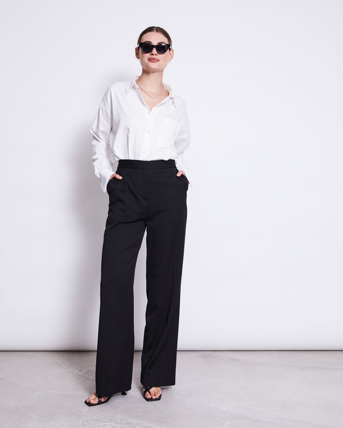 A person stands against a white background wearing the jan-n-june OVERSIZED SHIRT MAMRO WHITE, black sunglasses, wide-leg trousers, and heeled sandals, with their hands in their pockets.
