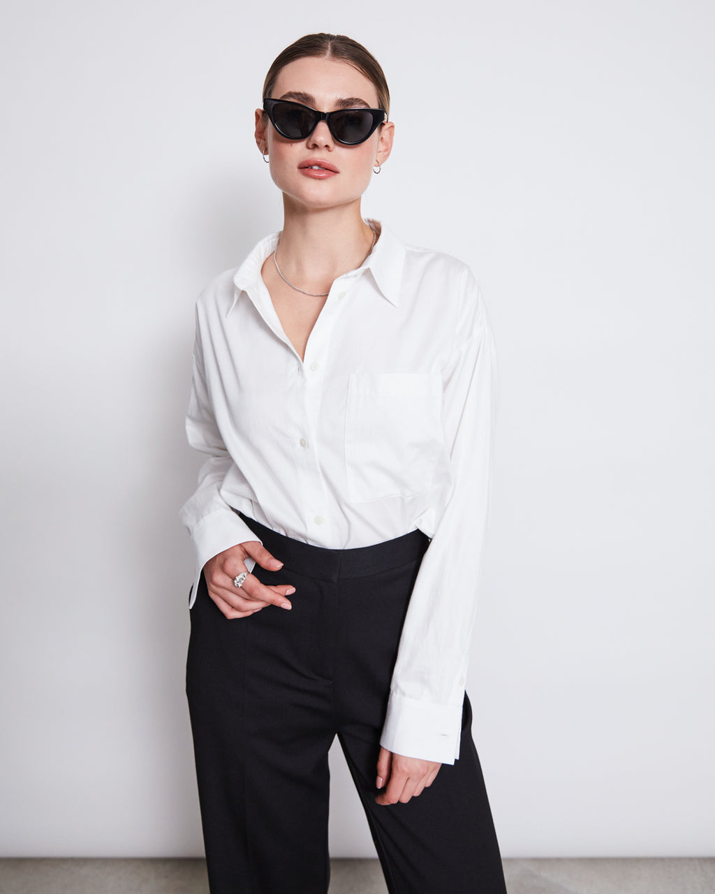 A woman with slicked-back hair wears the jan-n-june OVERSIZED SHIRT MAMRO WHITE, styled with black sunglasses and pants. She stands against a plain white background, one hand in her pocket.