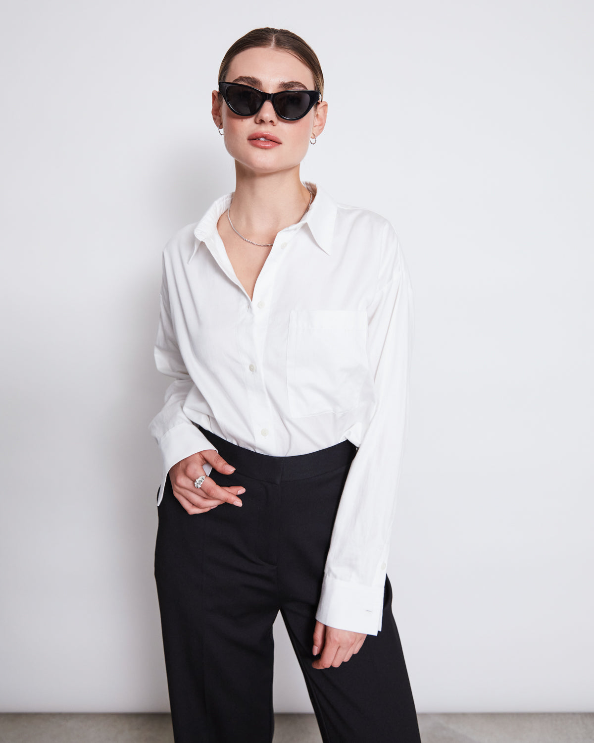 A woman with slicked-back hair wears the jan-n-june OVERSIZED SHIRT MAMRO WHITE, styled with black sunglasses and pants. She stands against a plain white background, one hand in her pocket.