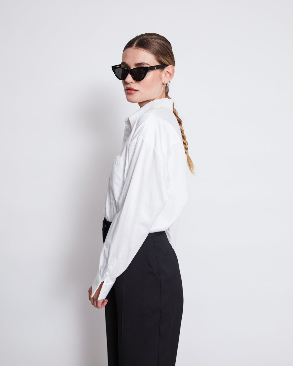 A woman with a long braid and black sunglasses wears the jan-n-june OVERSIZED SHIRT MAMRO WHITE, paired with high-waisted black pants, standing against a plain white background and looking to the side.