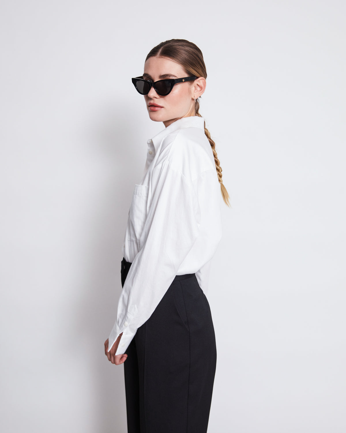 A woman with a long braid and black sunglasses wears the jan-n-june OVERSIZED SHIRT MAMRO WHITE, paired with high-waisted black pants, standing against a plain white background and looking to the side.