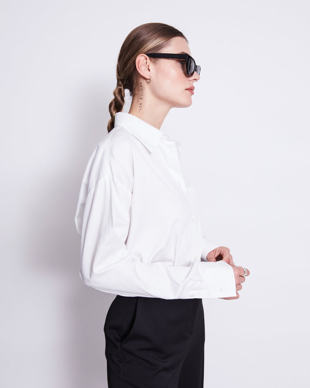 A woman with long brown hair in a braid wears black sunglasses, black pants, and the jan-n-june OVERSIZED SHIRT MAMRO WHITE, standing in profile against a plain white background.