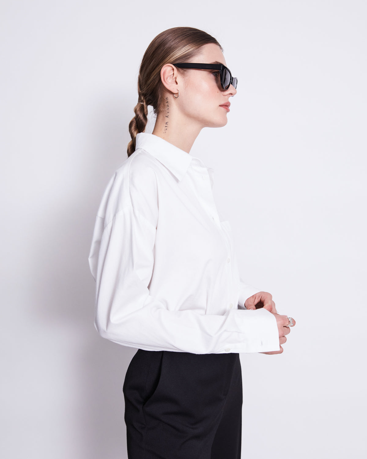 A woman with long brown hair in a braid wears black sunglasses, black pants, and the jan-n-june OVERSIZED SHIRT MAMRO WHITE, standing in profile against a plain white background.