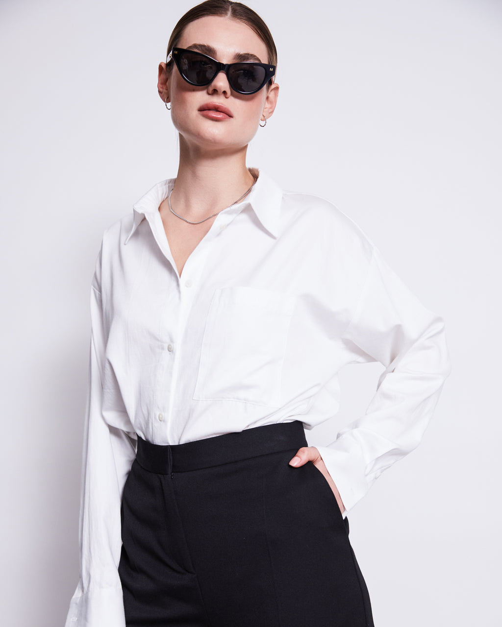 A person wearing black cat-eye sunglasses, the jan-n-june OVERSIZED SHIRT MAMRO WHITE, and high-waisted black pants stands confidently with one hand on their hip against a plain white background.