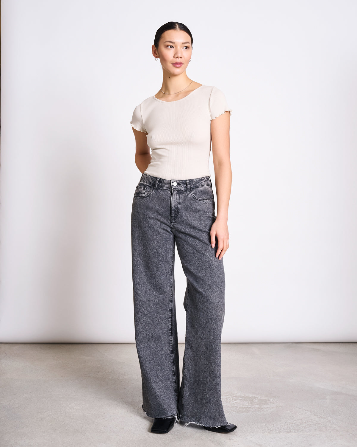 A woman with her hair tied back stands against a white background, wearing the jan-n-june MINIRIB T-SHIRT REN IVORY GOTS and high-waisted, wide-leg gray jeans with frayed hems. She completes the look with black shoes.