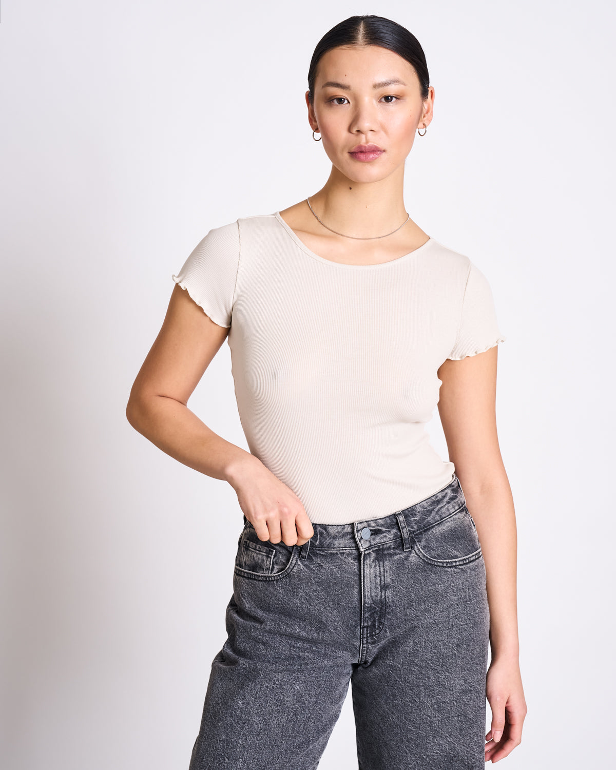 A person with straight dark hair pulled back wears the jan-n-june MINIRIB T-SHIRT REN IVORY GOTS and high-waisted black jeans, standing against a plain light background with one hand in their pocket.