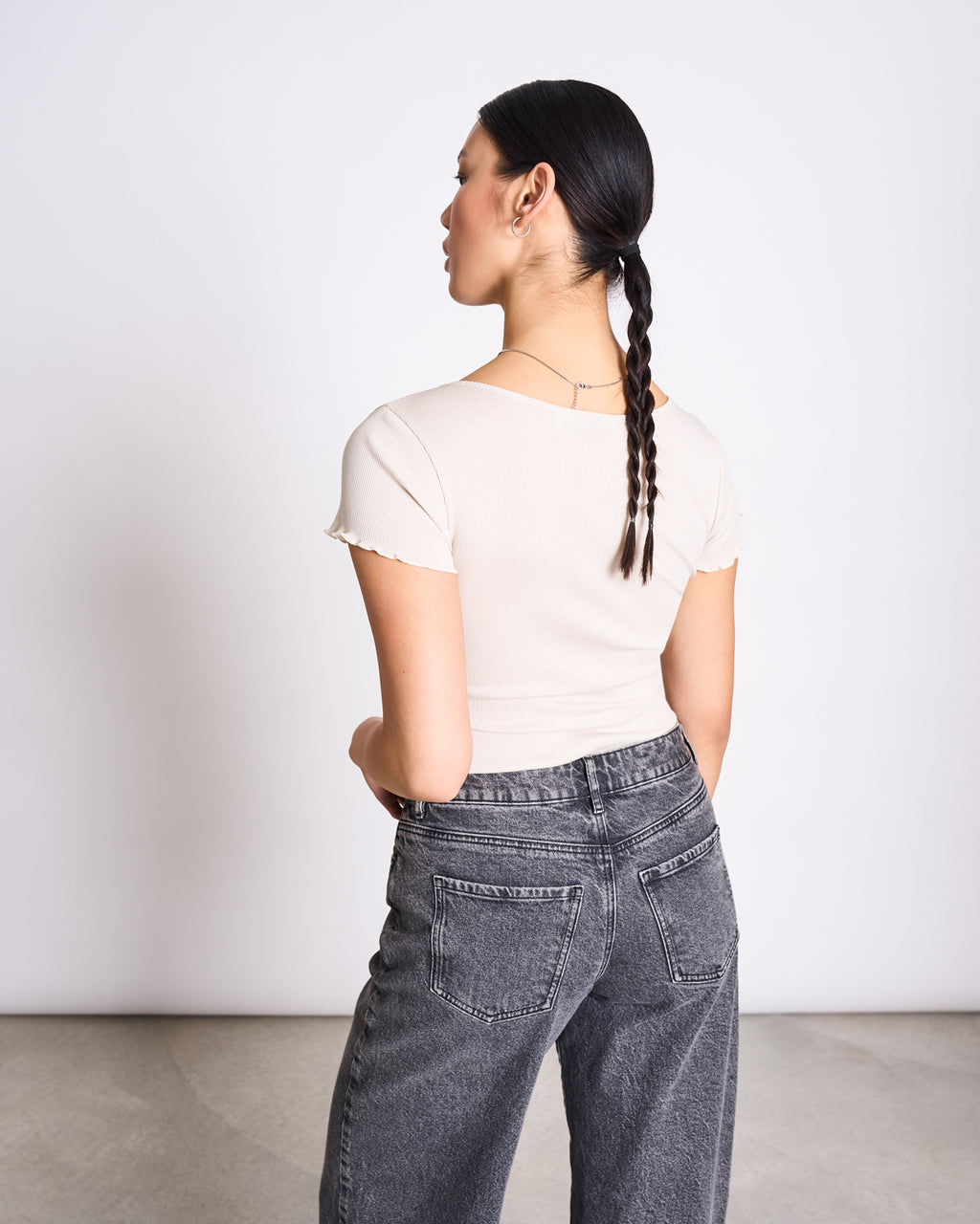 A woman with long dark braided hair, seen from behind in a minimalist indoor space, wears the jan-n-june MINIRIB T-SHIRT REN IVORY GOTS and high-waisted faded black jeans with scalloped hems.