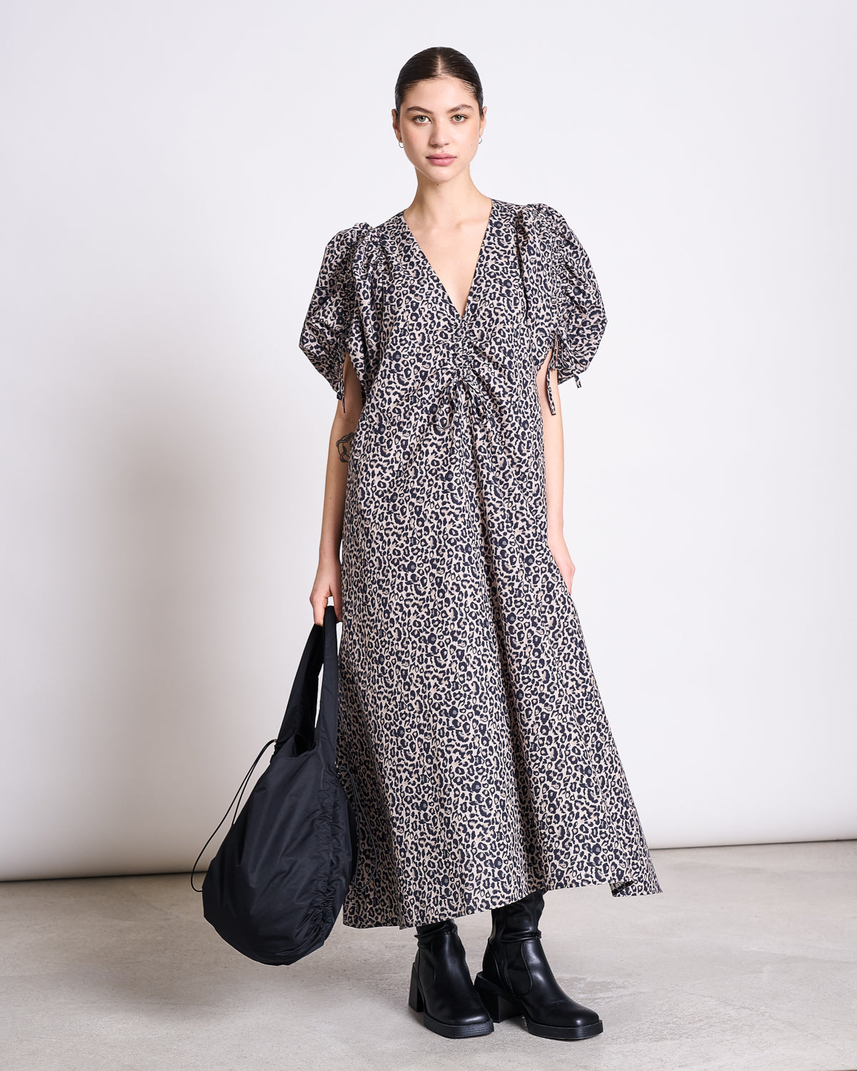 A woman stands against a plain light background, wearing the jan-n-june MAXI DRESS DANS LEO PRINT, styled with black ankle boots and a large black bag. Her hair is pulled back as she looks directly at the camera.