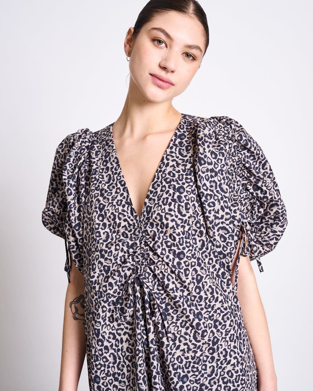 A woman with dark hair in a low bun wears the jan-n-june MAXI DRESS DANS LEO PRINT, an elegant v-neck maxi with a ruched front and short puffed sleeves in beige and black leopard print, standing against a plain white background.