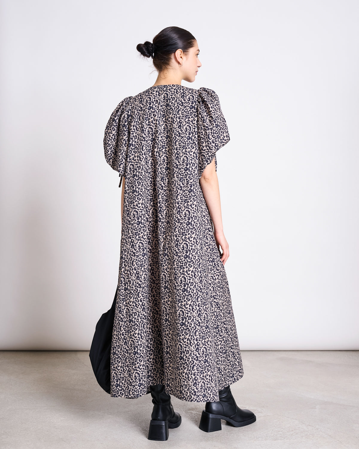A person with dark hair in a bun wears the jan-n-june MAXI DRESS DANS LEO PRINT, an ankle-length dress with puffed sleeves, paired with black chunky boots, standing indoors and facing away from the camera.