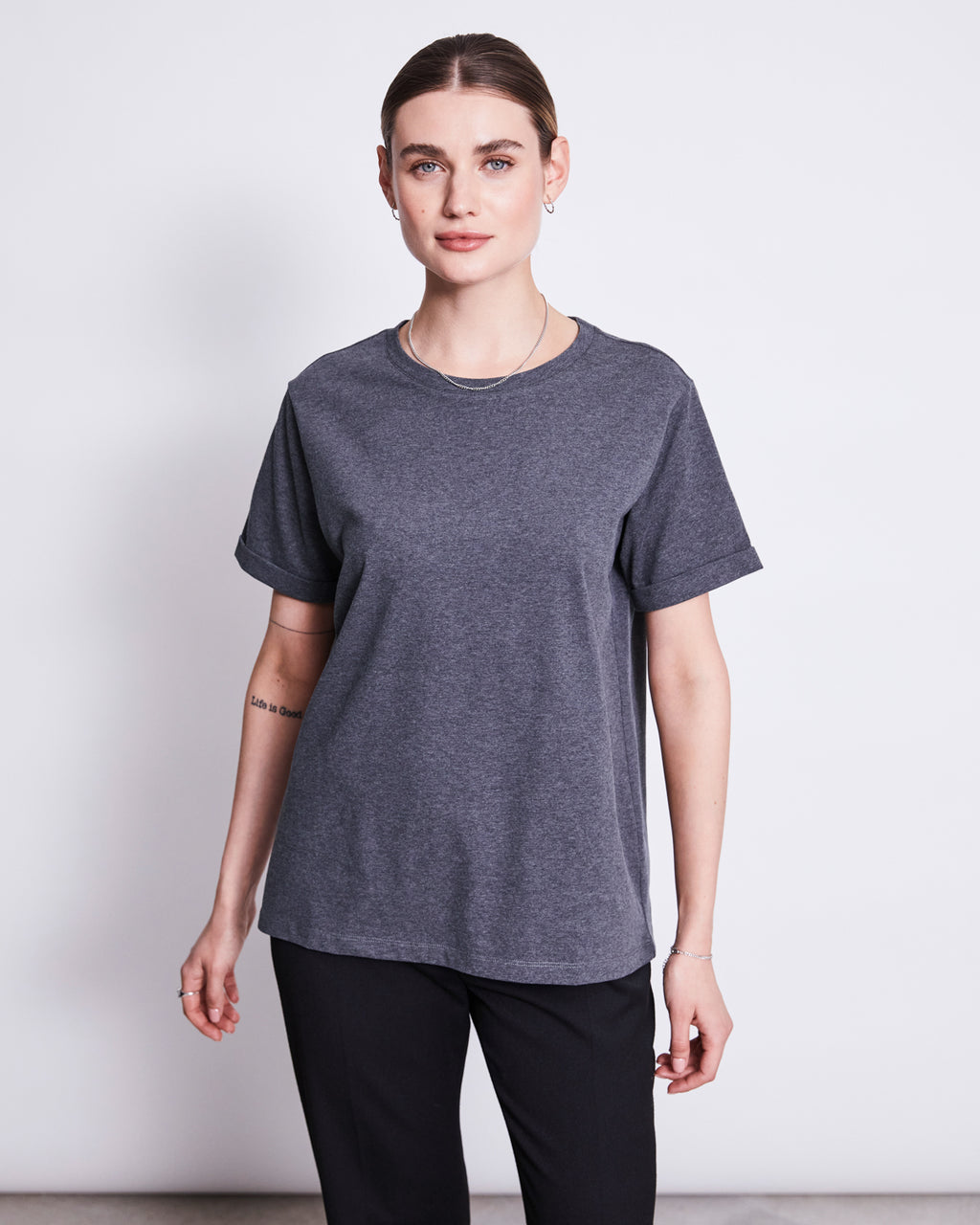 A woman with brown hair pulled back wears the jan-n-june BRUSHED T-SHIRT BOY DARK GREY GOTS and black pants, standing against a plain white background and looking at the camera with a neutral expression.