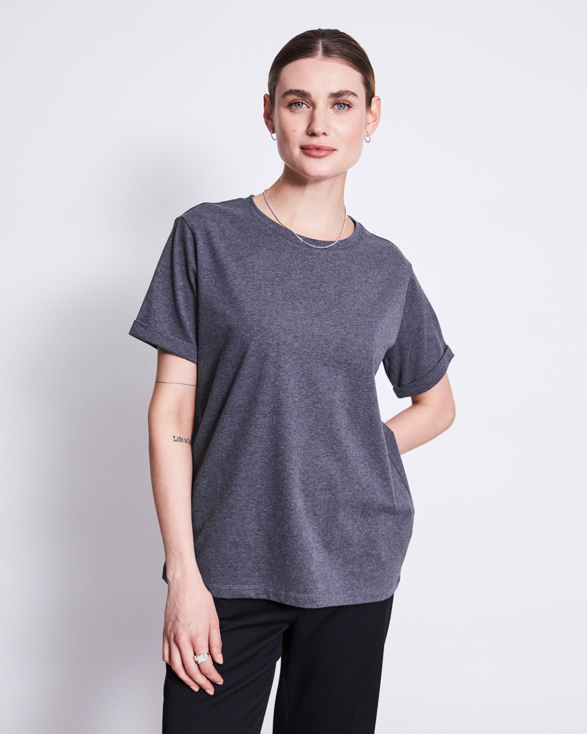A woman with light skin and brown hair tied back stands against a white background, wearing jan-n-junes BRUSHED T-SHIRT BOY DARK GREY GOTS in an oversized fit, paired with black pants, small hoop earrings, a necklace, and one hand in her pocket.
