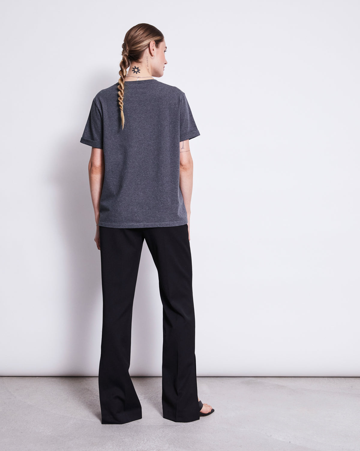A person with a long braid, wearing the jan-n-june BRUSHED T-SHIRT BOY DARK GREY GOTS and black pants, faces a white wall. A star tattoo is visible on their neck in a minimal setting with a plain floor and background.