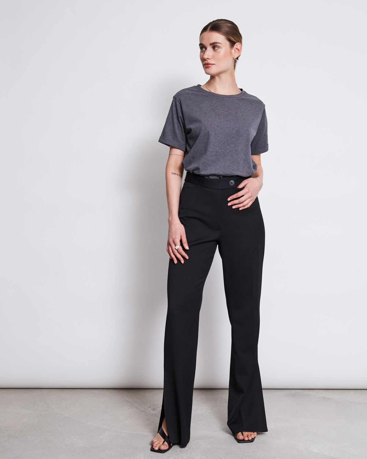 A woman, hair pulled back, stands by a white wall wearing the jan-n-june BRUSHED T-SHIRT BOY DARK GREY GOTS tucked into high-waisted black flared pants, looking to the side.