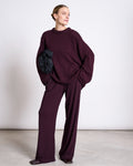 A woman stands against a plain background, wearing the jan-n-june JUMPER SONA CHERRY LACQUER GOTS—an oversized maroon knit sweater and matching wide-leg pants—holding a black ruched clutch. She looks to the side, smiling softly.