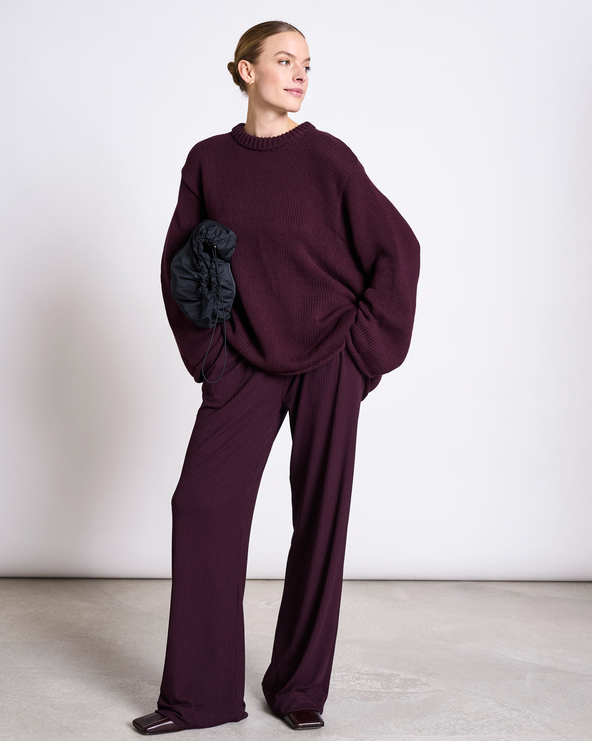 A woman stands against a plain background, wearing the jan-n-june JUMPER SONA CHERRY LACQUER GOTS—an oversized maroon knit sweater and matching wide-leg pants—holding a black ruched clutch. She looks to the side, smiling softly.