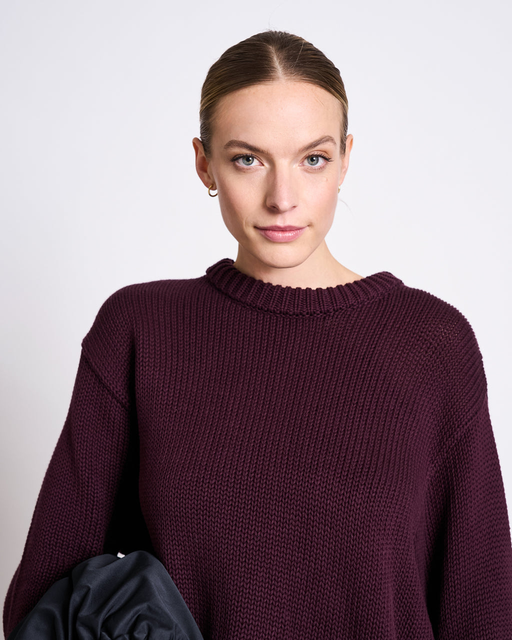 A woman with light skin and blonde hair wears the jan-n-june JUMPER SONA CHERRY LACQUER GOTS, an oversized maroon knit sweater, with small hoop earrings, standing against a plain white background and looking at the camera.