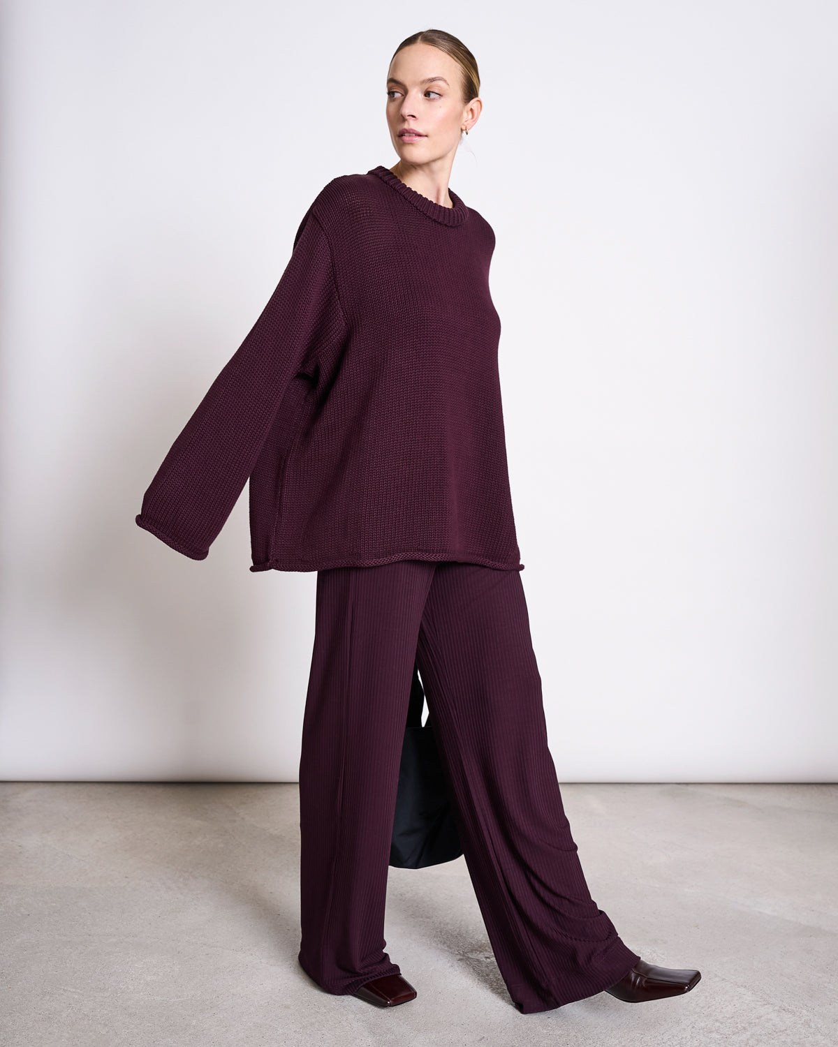 A person wearing jan-n-june’s LYOCELL RIB PANTS SMILLA CHERRY LACQUER and a relaxed burgundy sweater stands confidently against a white wall on a light floor, glancing to the side.