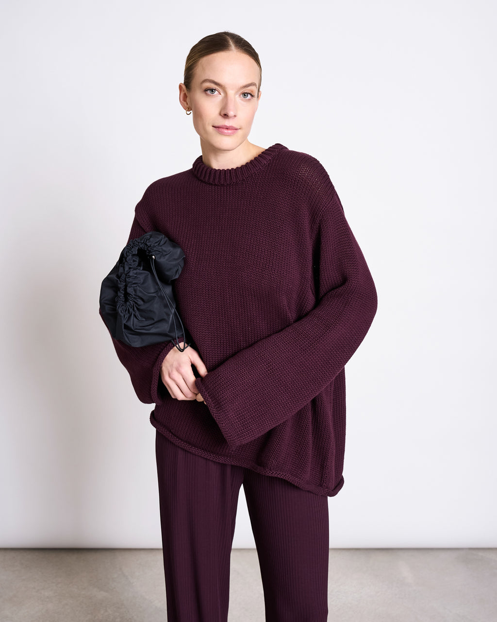 A woman stands against a plain white wall wearing the JUMPER SONA CHERRY LACQUER GOTS by jan-n-june—an oversized, dark burgundy organic cotton sweater with matching wide-leg pants—holding a black ruched clutch and gazing calmly at the camera.