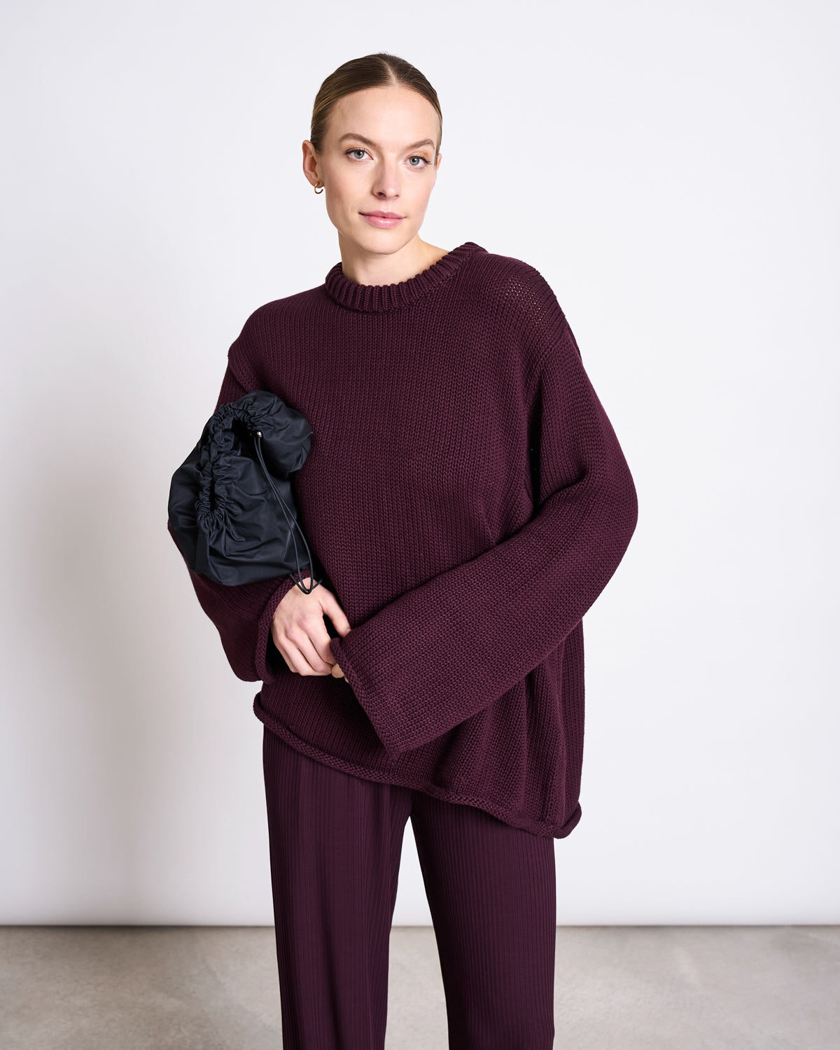 A woman stands against a plain white wall wearing the JUMPER SONA CHERRY LACQUER GOTS by jan-n-june—an oversized, dark burgundy organic cotton sweater with matching wide-leg pants—holding a black ruched clutch and gazing calmly at the camera.