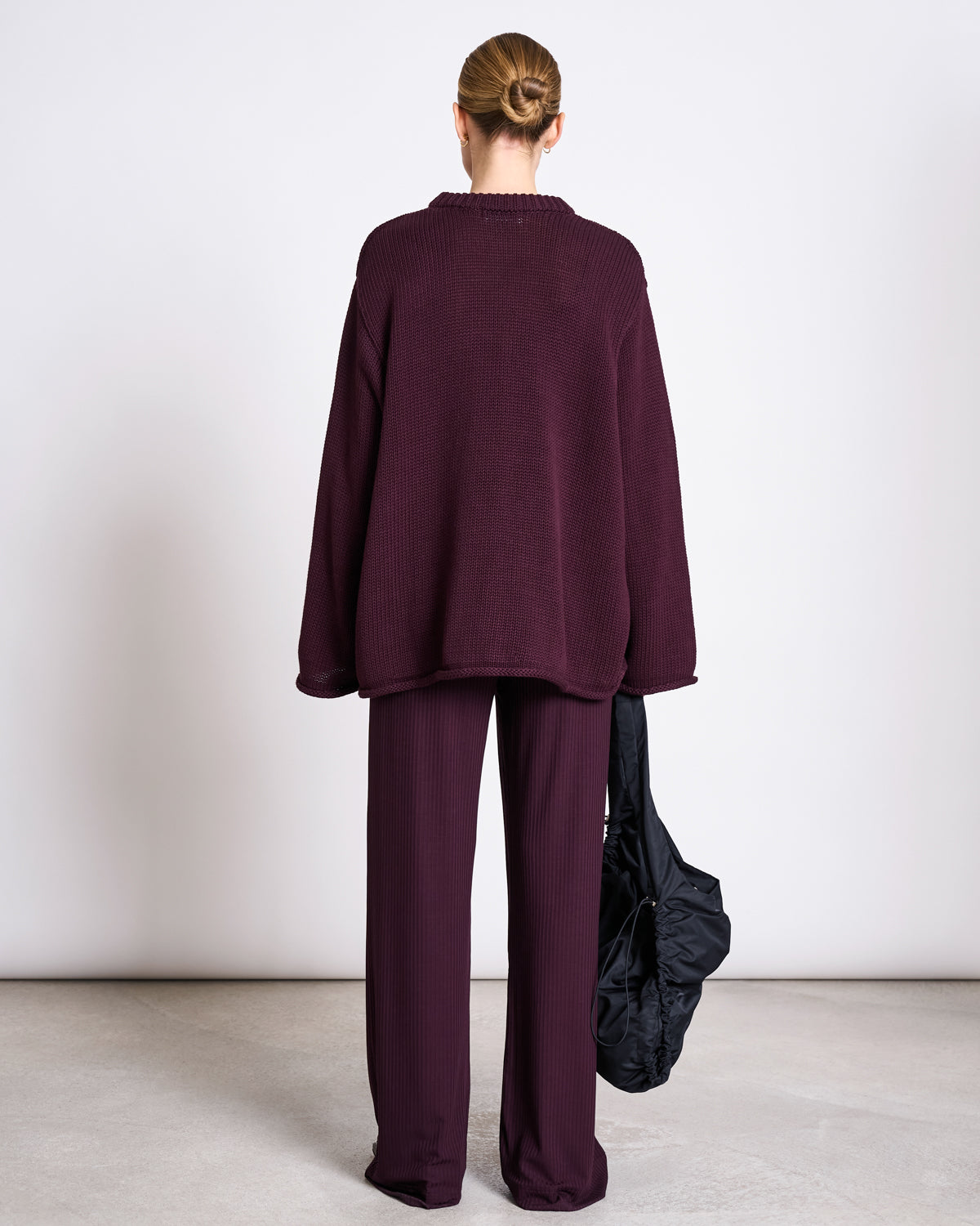 A person stands indoors, facing away from the camera, wearing jan-n-junes TENCEL™ LYOCELL RIB PANTS SMILLA CHERRY LACQUER and a matching dark sweater, holding a black bag with their hair styled in a neat bun.