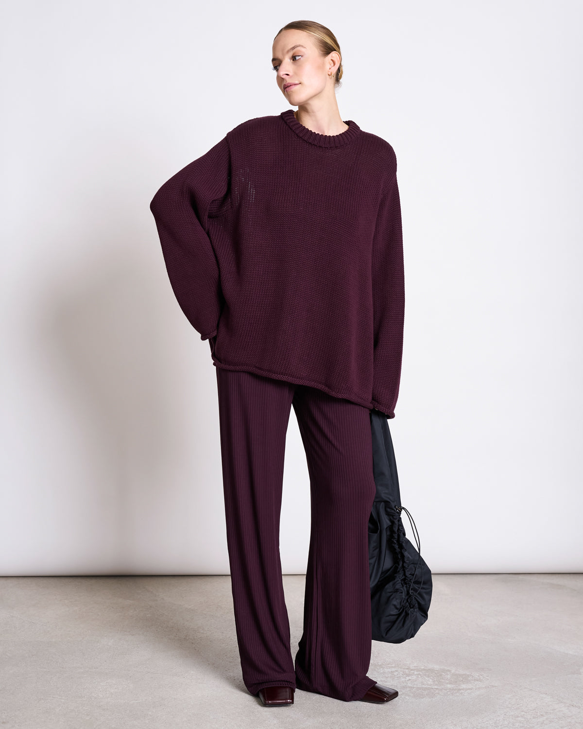 A person stands against a plain background wearing the jan-n-june JUMPER SONA CHERRY LACQUER GOTS, oversized and burgundy, with matching wide-leg pants, holding a black bag and looking off to their left.