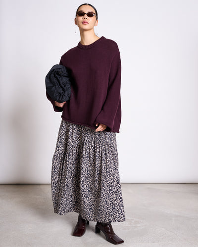 Wearing jan-n-junes JUMPER SONA CHERRY LACQUER GOTS, a person stands against a plain background, paired with sunglasses, a long leopard print skirt, dark boots, and holding a folded black garment under one arm.