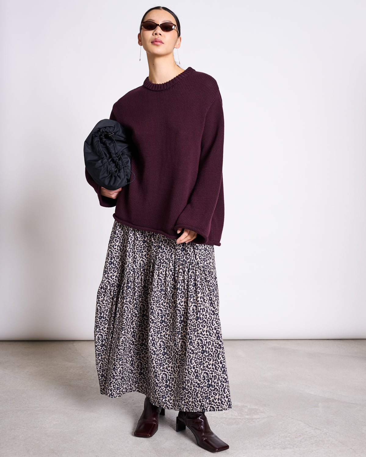 A person wearing sunglasses, a loose burgundy sweater, the jan-n-june TIERED MAXI SKIRT ALCA LEO PRINT, and dark ankle boots stands against a plain background, holding a black jacket in one arm.